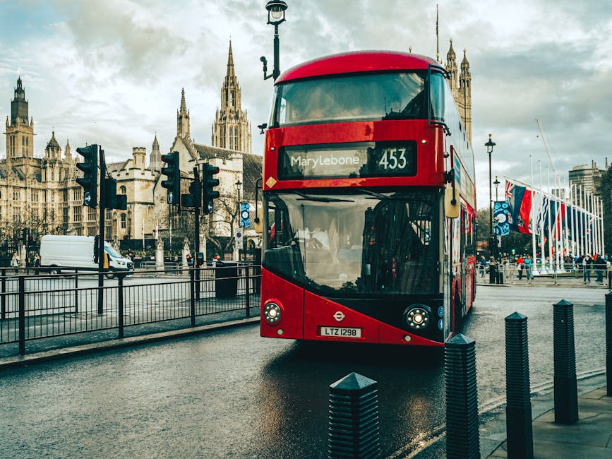 A red double-decker bus with route number 453 displaying 'Marylebone' as its destination, parked on a wet asphalt road near Marylebone Station in London. The bus features large glass windows, some reflecting the surrounding environment, and is situated in front of historic Gothic-style buildings with ornate spires and clock towers. The scene includes black bollards along the sidewalk, traffic lights, and a lamppost. On the right side, a row of British flags waves gently in the breeze. The area appears clean and well-maintained, with overcast skies casting a soft, diffused light that enhances the clarity of the scene. At Cleaner Marylebone, we specialise in surface cleaning and deep cleaning for both residential and commercial spaces, ensuring thorough sanitisation and hygiene that help keep areas like near Marylebone Station safe and spotless.