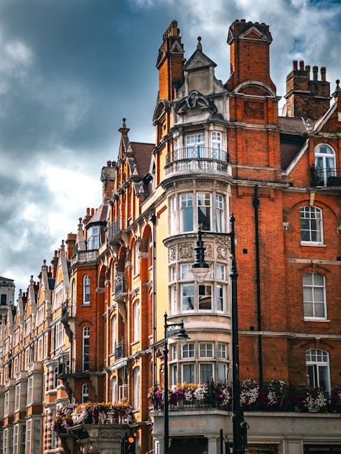 The image depicts a historic brick building in Marylebone under a partly cloudy sky. The building features ornate architectural details, including decorative stone trim around rectangular and arched sash windows, some with white frames. Several chimneys rise above the roofline, which is constructed of dark tiles. The ground-floor windows are complemented by flower boxes filled with colorful blooms, adding a touch of vibrancy to the structure. Street lamps are positioned along the sidewalk, and the overall scene captures the classic Victorian-era architecture typical of Marylebone. This exterior view emphasizes the charm and elegance of the area, aligning with the themes of thorough surface cleaning and maintenance promoted by [COMPANY_NAME] for residential or commercial properties in the district.