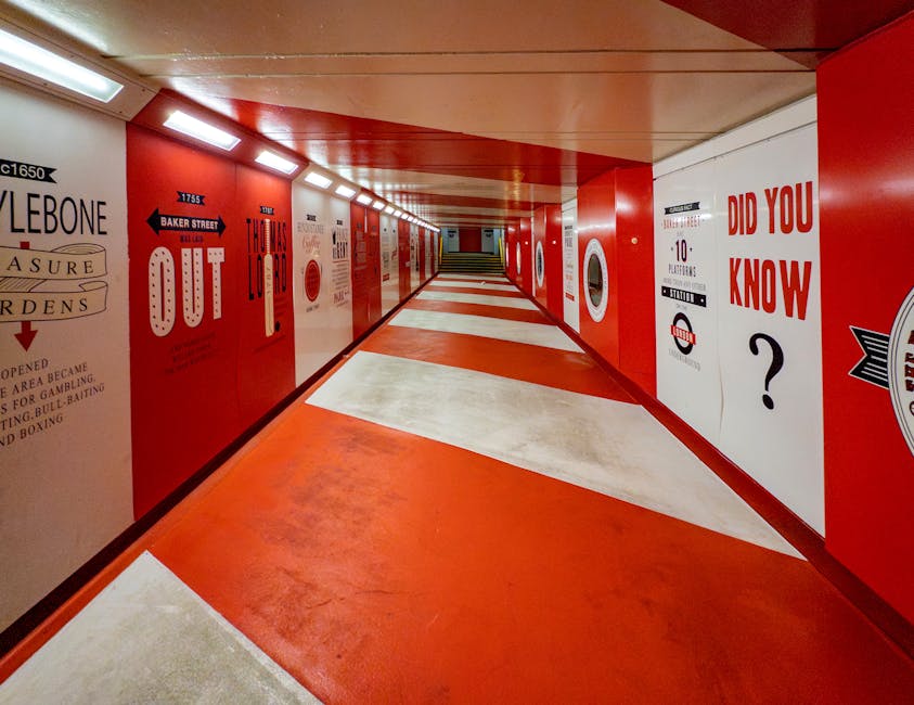 A brightly lit underground corridor with red and white walls decorated with posters and signs, including one that reads 'DID YOU KNOW?' and others with text and graphics promoting local events. The floor features a pattern of red and white tiles, some appearing slightly worn but overall clean. The ceiling has a series of rectangular ceiling lights, casting an even illumination throughout the space. The corridor is free of dust and marks, suggesting recent deep cleaning, which is aligned with the domestic cleaning and surface sanitisation services offered by Cleaner Marylebone. The setting exemplifies meticulous hygiene and maintenance of high-traffic areas, emphasizing the company's expertise in thorough cleaning processes.