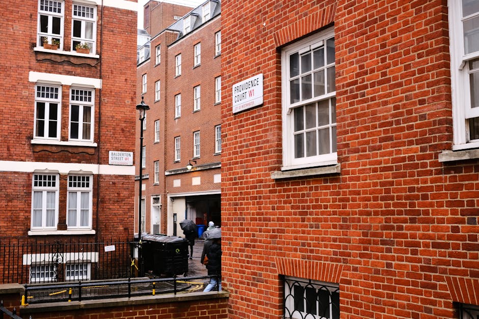 A street scene in Marylebone showing red-brick residential buildings with white-framed sash windows. The buildings feature traditional London architecture, with some windows adorned with window sills or decorative brick detailing. A narrow alleyway is visible between the buildings, with a black fence and a couple of pedestrians walking nearby. The scene is lit by natural daylight, highlighting the clean and well-maintained appearance of the area. The image depicts the external environment around a residential or commercial area in Marylebone, consistent with the standards of professional property maintenance and cleaning promoted by Cleaner Marylebone on their website.