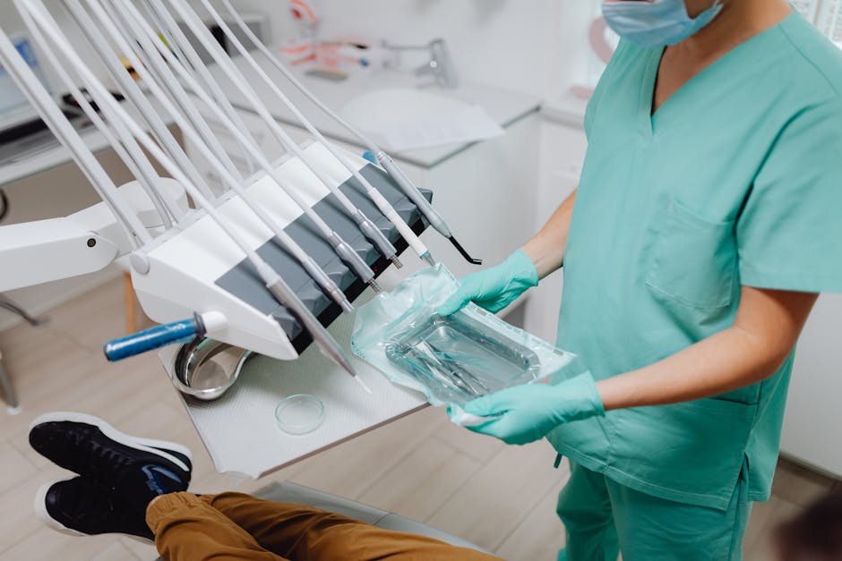 A healthcare professional dressed in light green medical scrubs and wearing a blue face mask and teal gloves is handling sterilized dental instruments in a clinical setting. The professional is holding a clear plastic packaging containing dental tools, preparing them for sterilization or safe use. On the light-colored examination table, there are additional tools and supplies, including a petri dish. In the background, there is a sterilization device with multiple orthodontic pliers and dental instruments organized. The environment is bright, clean, and sterile, with a focus on hygiene and surface cleanliness aligned with high standards for clinic sanitation, as promoted by Cleaner Marylebone for Harley Street clinic cleaning standards in Marylebone.