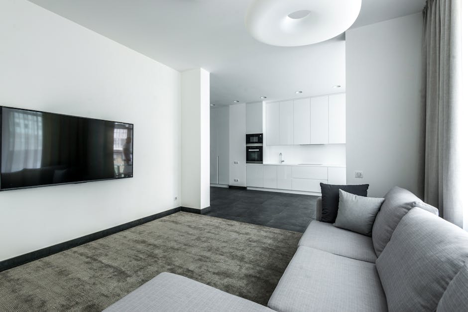 A modern, minimalist living room with a grey fabric sofa adorned with black and grey cushions, placed on a beige carpet. A wall-mounted flat-screen TV is positioned on a white wall to the left. The room is filled with natural light from a large window with light grey curtains on the right side. The open-plan space extends into a sleek, white kitchen with built-in appliances, including an oven and microwave, and a white countertop with a stainless steel sink. The floors transition from carpeted in the living area to dark grey tiles in the kitchen, and the room has a clean, clutter-free appearance consistent with professional domestic cleaning standards promoted by Cleaner Marylebone for deep cleaning and surface sanitisation.