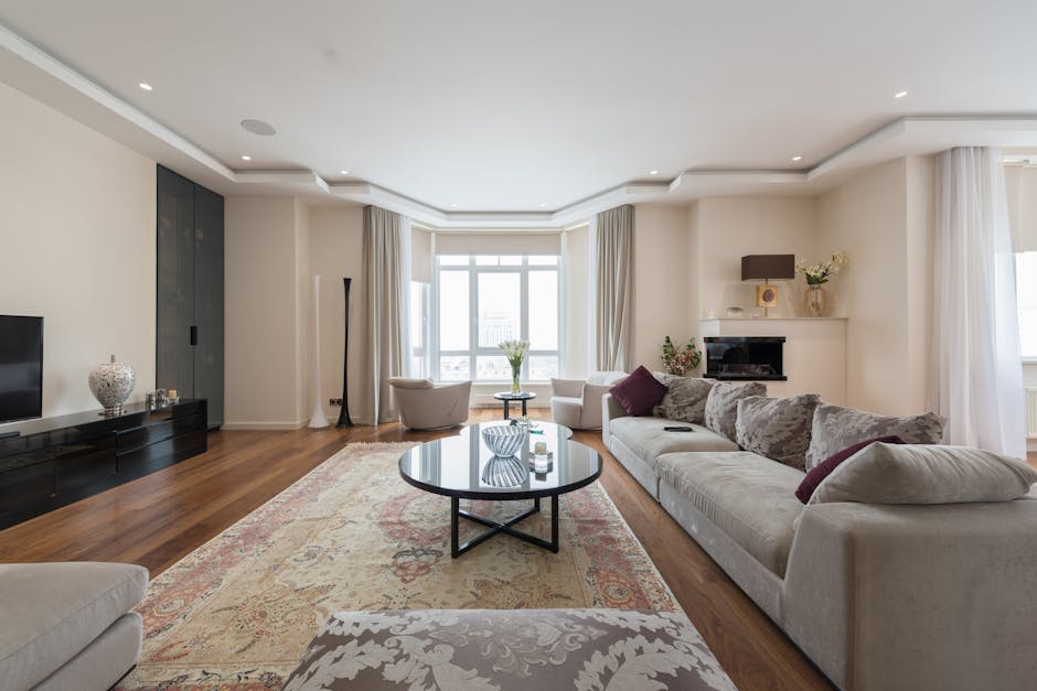 A spacious, well-lit living room featuring a large cream-colored sofa with purple and beige throw pillows, a patterned area rug covering a polished wooden floor, and a round glass coffee table with decorative items. The room has a modern design with white walls, a flat-screen TV on a black entertainment unit, and large windows draped with white curtains allowing natural light to fill the space. A fireplace with a small plant and artwork above it is visible on one wall, and the room appears clean, tidy, and maintained with no visible dust or clutter. Cleaner Marylebone offers surface cleaning and deep cleaning services to ensure such spaces remain hygienic and odour-free.