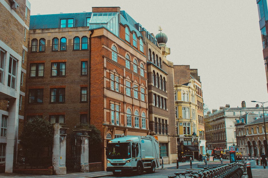 A street scene in Marylebone showing a row of multi-story buildings with various architectural styles, including brick and stucco facades, large windows, and decorative elements. The street features a white waste collection vehicle parked near the curb, with bollards and bicycles lined up along the sidewalk. The area is well-lit with natural daylight, and the street appears clean and tidy, reflecting effective rubbish removal and maintenance by Cleaner Marylebone. The scene captures the urban environment with a focus on surface cleanliness and the neat presentation of the surroundings, typical of thorough domestic or commercial cleaning and waste management services.