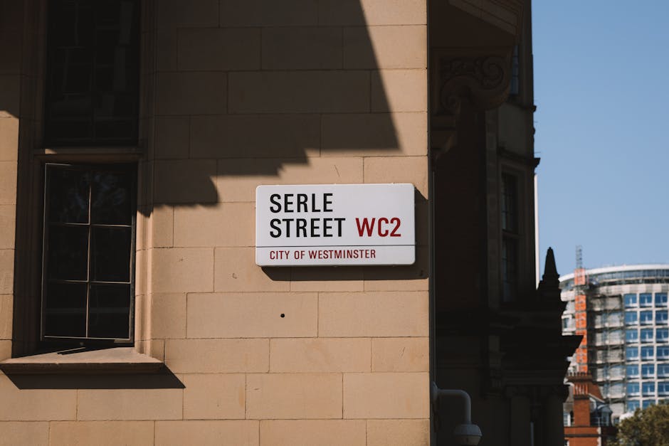 A street sign on the beige stone exterior wall of a building at Serle Street, WC2, in Westminster, London. The sign displays the street name in black text on a white background with the postcode 'WC2' in red and the label 'City of Westminster' below. Adjacent to the sign is a window with black framing, reflecting some sky and surroundings. The scene is illuminated by natural daylight, casting shadows on the wall, and in the background, there are modern high-rise buildings with glass facades under a clear blue sky. This image relates to urban street signage, with the clean and well-maintained building exterior visible for context, as part of the local district in Westminster, London.