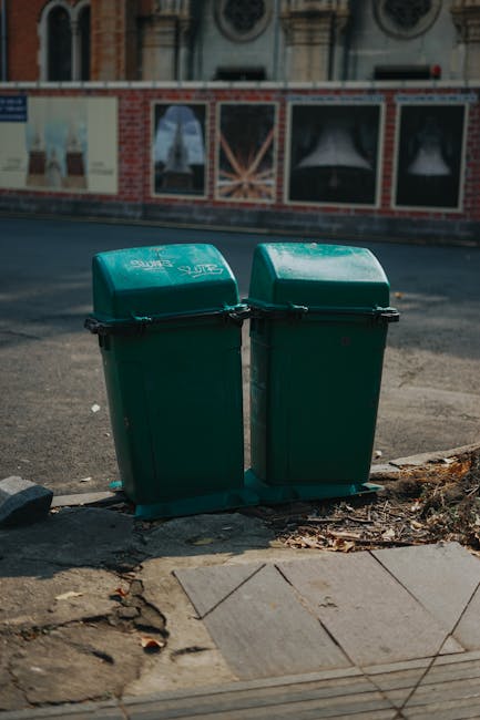 Two green plastic waste bins with black handles and compact lids are positioned on a concrete sidewalk in front of a street, adjacent to a crack in the pavement and some debris. The bins appear clean and are situated near a curb that separates the sidewalk from the roadway. Behind the bins, there is a brick and boarded-up window wall with three large darkly tinted windows covered with newspaper, suggesting a building under renovation or construction. The lighting is natural daylight, illuminating the scene clearly. This setting exemplifies basic waste management and cleanliness practices, aligning with the importance of proper disposal and hygiene in residential or commercial environments, such as those maintained by Cleaner Marylebone, especially within the context of Westminster Council waste rules for tenants.